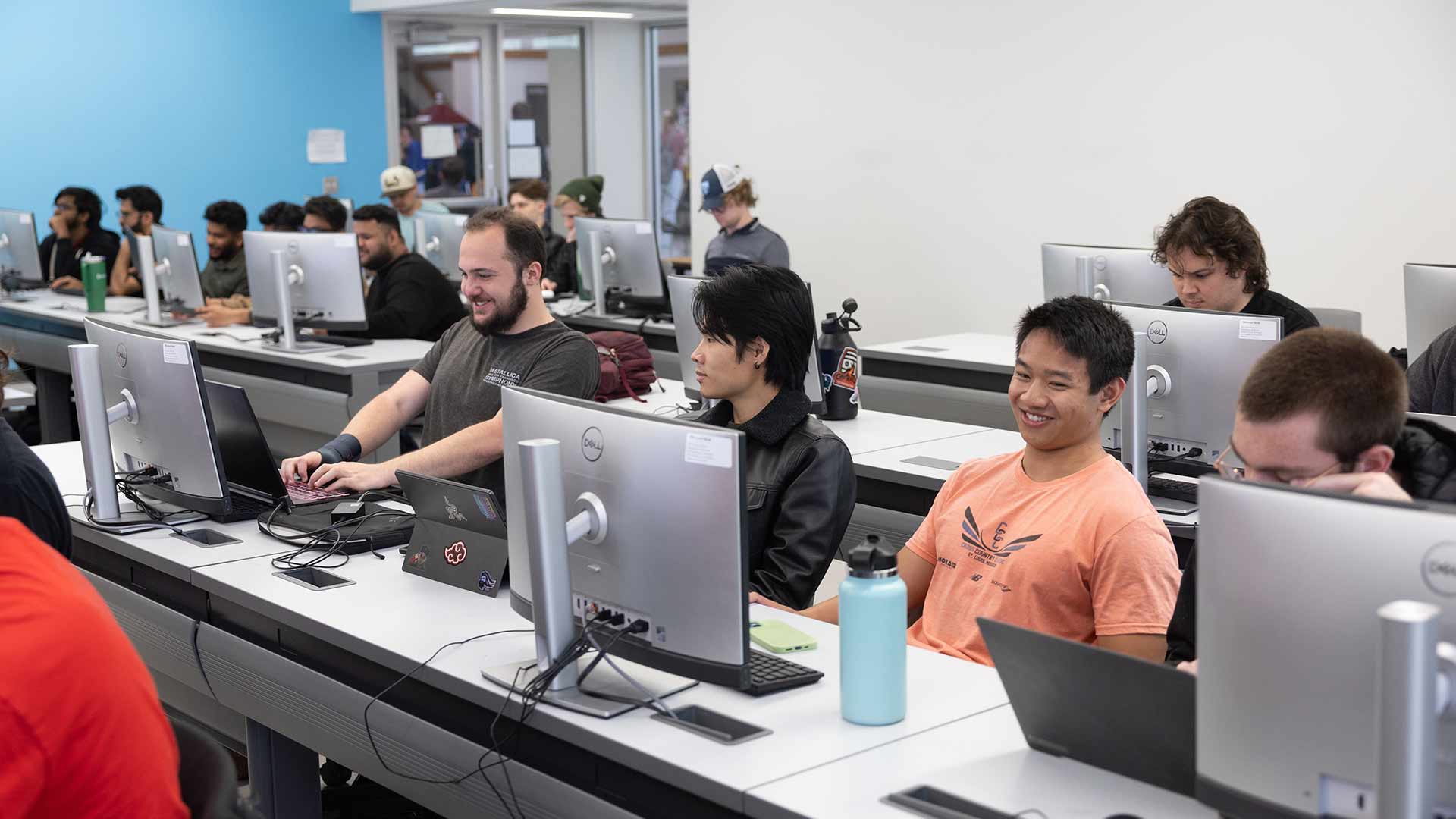 Three computer science students smile while working on a class project during an Intro to Software Development (CSC 450) course. Other students are seen in the background.