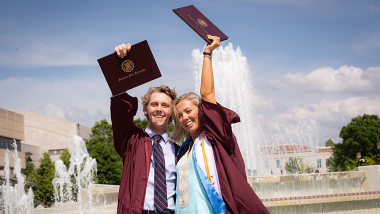 Two Missouri State graduates holding up their diploma covers in front of the campus fountain.