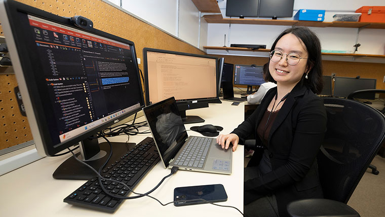 A computer science student smiles for a photo as she performs research on her laptop and desktop computers.