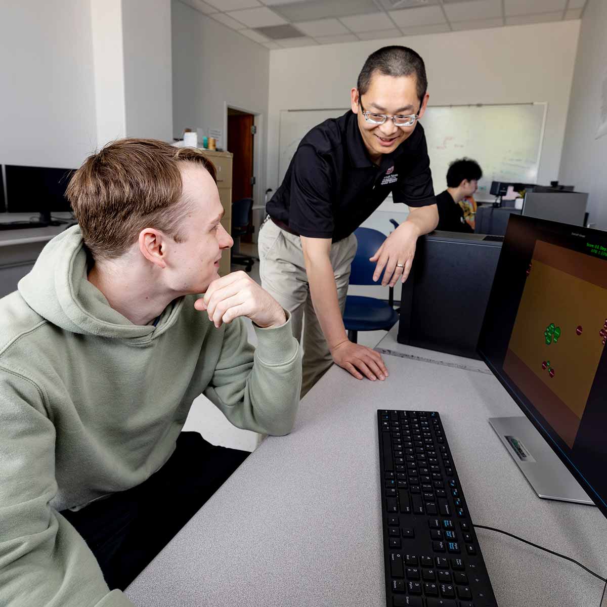 Dr. Siming Liu and a graduate student share a laugh while working together in a research lab. Blocks of data are shown on a computer monitor in front of them.