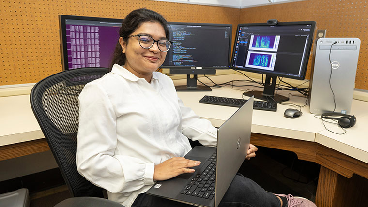 A computer science student smiles at the camera while studying in a research lab on campus. Three computer monitors displaying data are seen in the background.