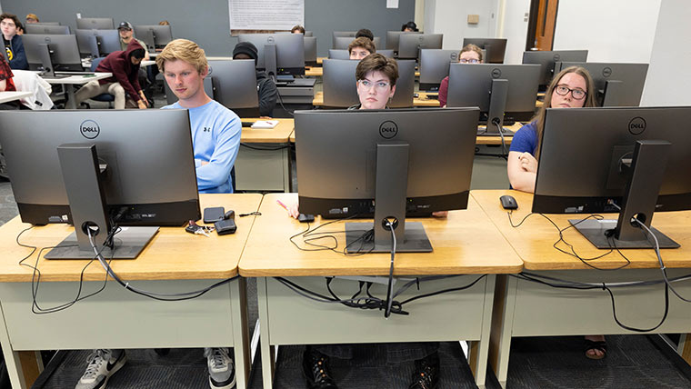 Three computer science students seated in the front row of a class at their computer stations.