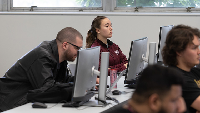 A computer science student seated at her computer station and wearing Bearwear listens attentively during class.